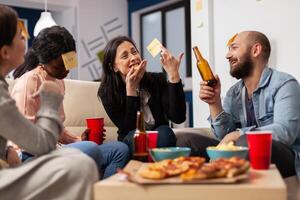 Cheerful group of colleagues enjoying a game of charades after work at office. Multi ethnic people play imitation concept for fun activity entertainment while eating pizza and drinking beer photo
