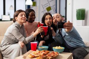 Diverrse group of coworkers taking selfie on smartphone after work at office party. Cheerful colleagues having fun at celebration meting with pizza chips bottles and cups of beer alcohol photo
