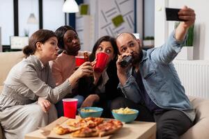 Multi ethnic team of friends make memories at after work party taking pictures on smartphone. Coworkers celebrating at office space with snacks pizza chips cups of drinks on table photo