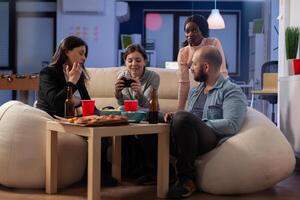Multi ethnic group of coworkers play game on console while holding joystick. Cheerful diverse friends enjoy fun competition on tv computer while having snacks and beer of table photo