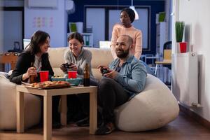Diverse team of workers playing console game on tv at office after work using joysticks on sofa. Multi ethnic friends starting competition while having snacks pizza chips and drinking beer photo