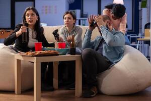 Team of multi ethnic friends losing tv game on console after work at office. Cheerful diverse group play with joystick while eating snacks pizza chips and drinking beer alcohol from cups photo