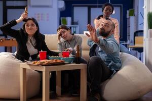 Team of multi ethnic friends losing tv game on console after work at office. Cheerful diverse group play with joystick while eating snacks pizza chips and drinking beer alcohol from cups photo