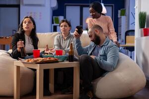 Diverse group of coworkers play cheerful game on tv console while holding controller joystick. Multi ethnic friends having fun entertainment at party with snacks and cups of drinks photo