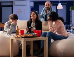 Diverse group of mates bonding while playing game on tv console losing with joystick controller after work. Multi ethnic team enjoy office celebration party with snacks and drinks photo