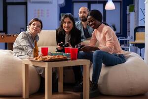 Multi ethnic diverse group of friends playing fun game on tv console after work at office. Cheerful colleagues holding joystick controller for gaming indoors on sofa. Happy team celebrating photo