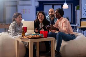 Multi ethnic diverse group of friends playing fun game on tv console after work at office. Cheerful colleagues holding joystick controller for gaming indoors on sofa. Happy team celebrating photo