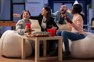 Multi ethnic friends celebrate together after work at office while playing game on tv console with controller joysticks. Diverse group of colleagues having fun gaming at party celebration photo