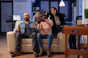 Group of multi ethnic friends using vr glasses for fun after work at office while holding controller joystick. Diverse team enjoy celebration party bonding together with drinks and snacks photo