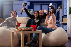 Multi ethnic friends celebrate together after work at office while playing game on tv console with controller joysticks. Diverse group of colleagues having fun gaming at party celebration photo