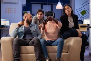 Group of multi ethnic friends using vr glasses for fun after work at office while holding controller joystick. Diverse team enjoy celebration party bonding together with drinks and snacks photo