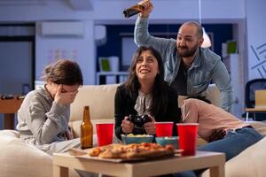 Diverse group of mates bonding while playing game on tv console losing with joystick controller after work. Multi ethnic team enjoy office celebration party with snacks and drinks photo