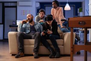 Multi ethnic team of mates play with vr glasses after work at office party. Diverse group of friends enjoy gaming experience with console controller joystick for victory celebration photo