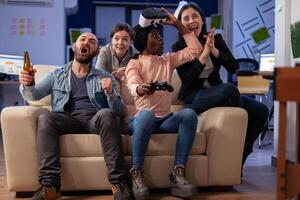 Diverse team of colleagues enjoy vr glasses experience after work at office while using controller joystick. Multi ethnic friends bonding and celebrating relaxation fun enjoyment photo