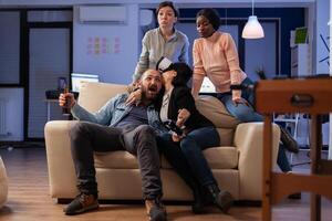Multi ethnic team of mates play with vr glasses after work at office party. Diverse group of friends enjoy gaming experience with console controller joystick for victory celebration photo