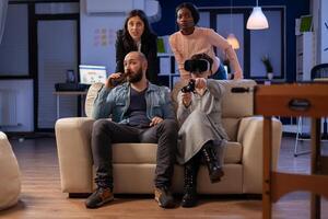 Cheerful multi ethnic friends enjoy vr glasses experience after work at office party. Diverse people play with controller joystick on tv console having fun together bonding at celebration photo