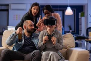 Diverse team of people use vr glasses on tv console at office party celebration after work. Multi ethnic colleagues play with joystick controller having fun enjoyment entertainment photo