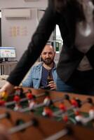 Portrait of business man drinking beer from bottle at office party after hours, enjoying leisure activity with colleagues. Cheerful adult having fun with after work drinks and foosball game. photo