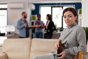 Portrait of young woman holding beer bottle after hours in office, celebrating with workmates. Happy person enjoying leisure activity and party after work, having alcoholic drinks. photo