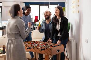 Diverse group of colleagues having fun with foosball table game, enjoying after work drinks and entertainment activity. Office coworkers celebrating after hours with alcohol and pizza. photo