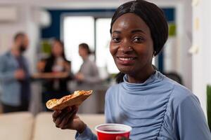 Portrait of african american woman holding pizza and beer, enjoying after work drinks party with office coworkers. Cheerful person having fun at celebration after hours with alcohol and snacks. photo