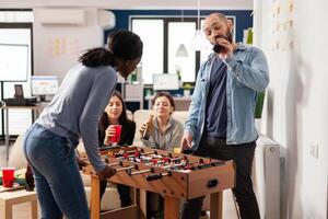 Multiethnic group of people playing at foosball table game, meeting for drinks in office after work hours. Man and women drinking beer from bottles and eating snacks at celebration. photo