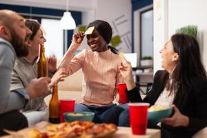 African american woman playing guess who game with coworkers, celebrating with drinks after work. Office colleagues enjoying charades pantomime play with sticky notes at party. photo