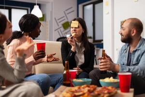 Multiethnic group of friends using sticky notes to play guess who game together at office party after hours. Business people playing charades, eating pizza and drinking beer after work. photo