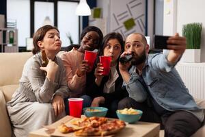 Group of people taking selfies and celebrating with drinks in office after work hours. Man and women having fun with pictures on smartphone, enjoying alcoholic drinks and snacks at party. photo