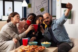 Diverse group of colleagues using mobile phone to take pictures at office party with drinks after hours. People doing fun leisure activity with smartphone, having beer bottles and pizza. photo