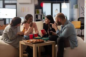 Multiethnic group of coworkers having conversation at office celebration. Man and women talking and having fun with alcoholic drinks, pizza and chips at party after work hours. photo