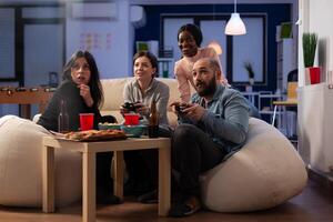 Smiling people playing games online on tv console, using controller to win at office part after work. Colleagues having fun with game, eating pizza and drinking alcoholic beer. photo