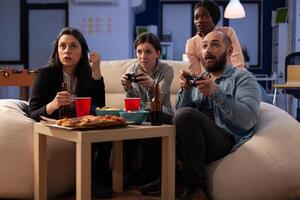 Office colleagues enjoying games play with joystick on tv console at drinks party after hours. Cheerful friends having fun with technology, alcoholic beverage and snacks after work. photo