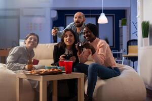 Diverse group of colleagues playing games on tv console with controller, enjoying leisure activity with drinks after work hours. People gaming, drinking beer and eating pizza. photo