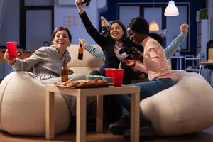 Multiethnic group of people winning games play with joystick on console, meeting in office for drinks after work hours. Man and women celebrating game win and drinking beer. photo