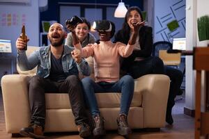 Diverse group of workmates playing games with vr headset, enjoying virtual reality technology on game play with joystick. Cheerful people having fun at office party after work. photo