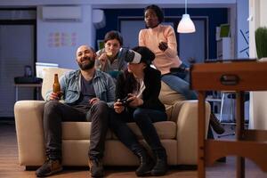 Multiethnic group of people having fun with games on console and virtual reality glasses. Coworkers meeting at office party celebration with beer drinks after work hours. photo