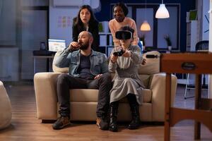 Multiethnic group of workmates having fun with videogames and virtual reality glasses at drinks party in office. People enjoying vr technology and online play at celebration after work. photo