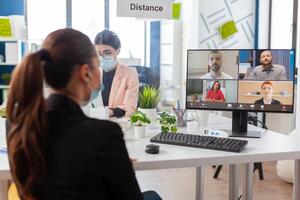 Back view of woman talking with team during remote conference in new normal office in timp of global pandemic with coronavirus, keeping social distancing wearing face mask. photo