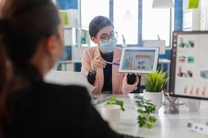 Close up of coworkers with face mask working together at financial project using tablet computer while sitting in company office. Team mantain social distance to avoid infection with covid19. photo