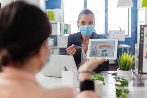Back shot of business womens with medical face mask working together at management presentation using tablet computer while sitting in company office. Team respecting social distance. photo