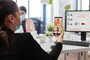 Businesswoman holding smartphone discussing with remote friends having online videocall conference meeting working in company office. Manager with face mask against covid19 during teleconference photo