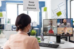 Businesswoman with medical face mask discussing marketing statistics with remote teamwork having online videocall meeting conference on laptop working in startup office. Teleconference on screen photo