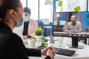 Businesswoman with medical face mask against covid19 discussing financial strategy with remote manager during online videocall meeting conference in startup office. Teleconference on computer screen photo