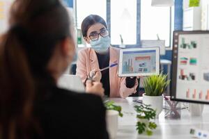 Businesswoman wearing medical face mask against covid19 showing company graph to collegue using tablet computer working in startup office. Teamwork planning management strategy during global pandemic photo