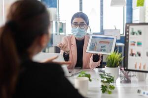 Businesswoman wearing medical face mask against covid19 showing financial graph to collegue using tablet computer working in startup office. Teamwork planning marketing strategy during global pandemic photo