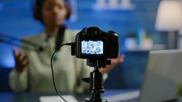 Close up of professional camera sitting in front of dslr recording online show. African woman vlogger speaking at microphone during livestreaming, blogger discussing in podcast wearing headphones photo