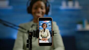Close up of smartphone recording vlog of african influencer in home studio using smartphone. Speaking during livestreaming, blogger discussing in podcast wearing headphones and professional microphone photo