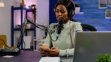 African vlogger recording blog reading messages from smartphone with modern equipment in studio podcast. Influencer looking at phone talking in broadcast using tech to connect with audiences photo