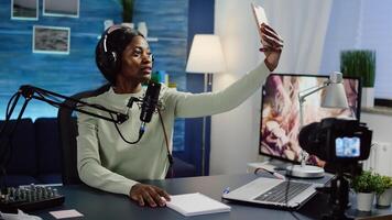 Black woman vlogger taking selfie using smartphone during streaming live content for social media. Influencer speaking during livestreaming, blogger discussing in podcast wearing headphones. photo